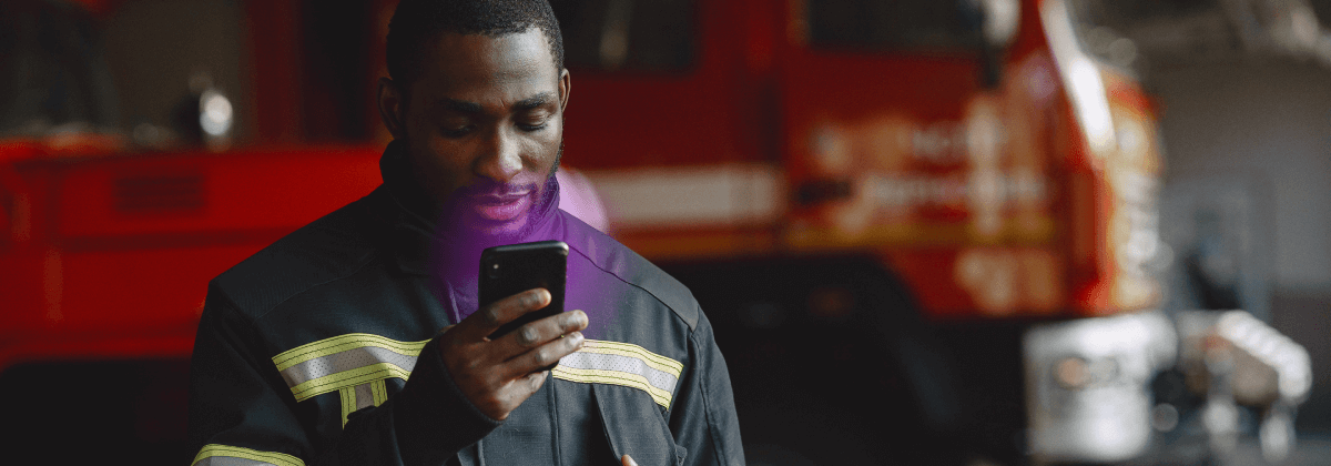 Joven bombero mirando un teléfono móvil del que sale un resplandor púrpura. En el fondo se ve un camión de bomberos.
